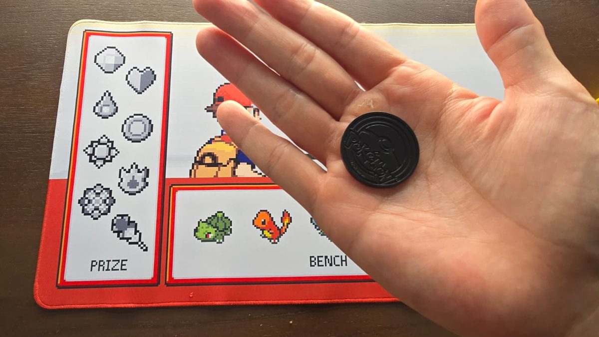 A black Pokémon TCG coin showing the Poké Ball logo side, held in a palm